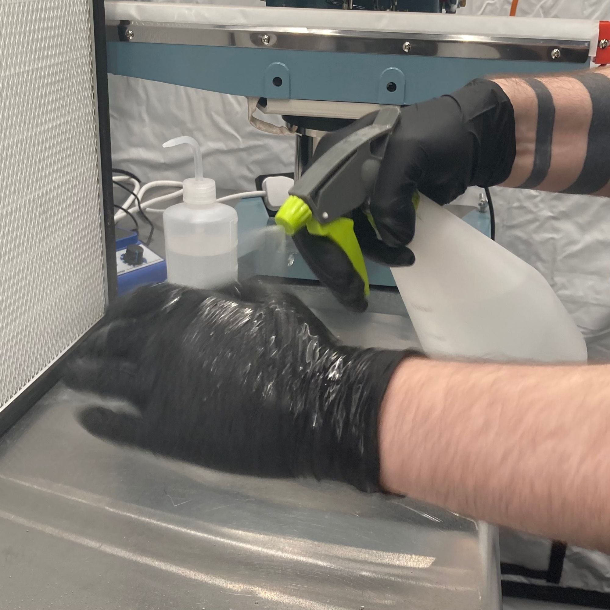 Person wearing black gloves using a spray bottle on a surface with a machine in the background.